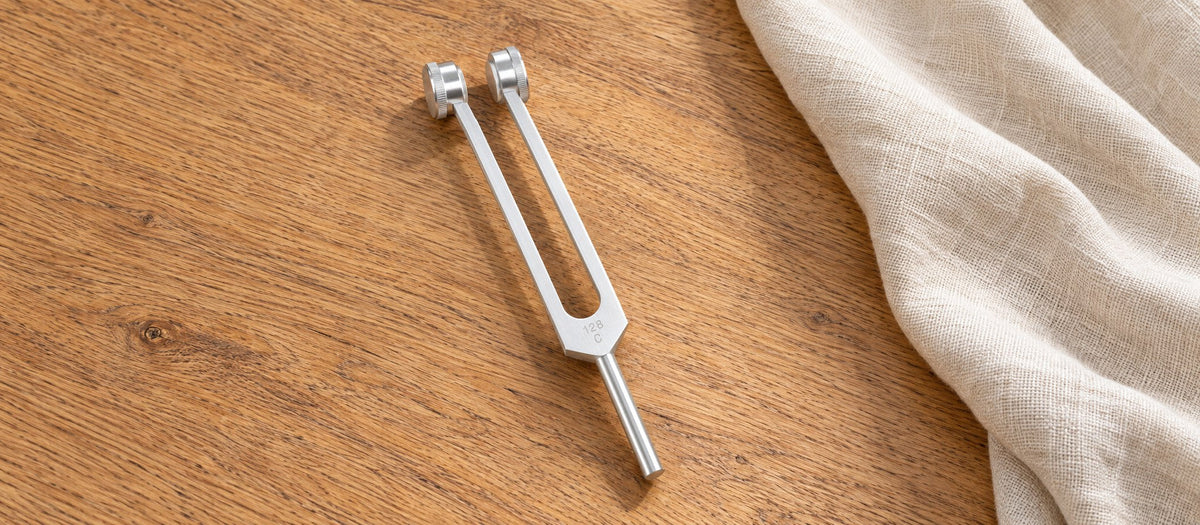 Weighted 128 C tuning fork with paired cylindrical weights on an oak table beside ivory linen