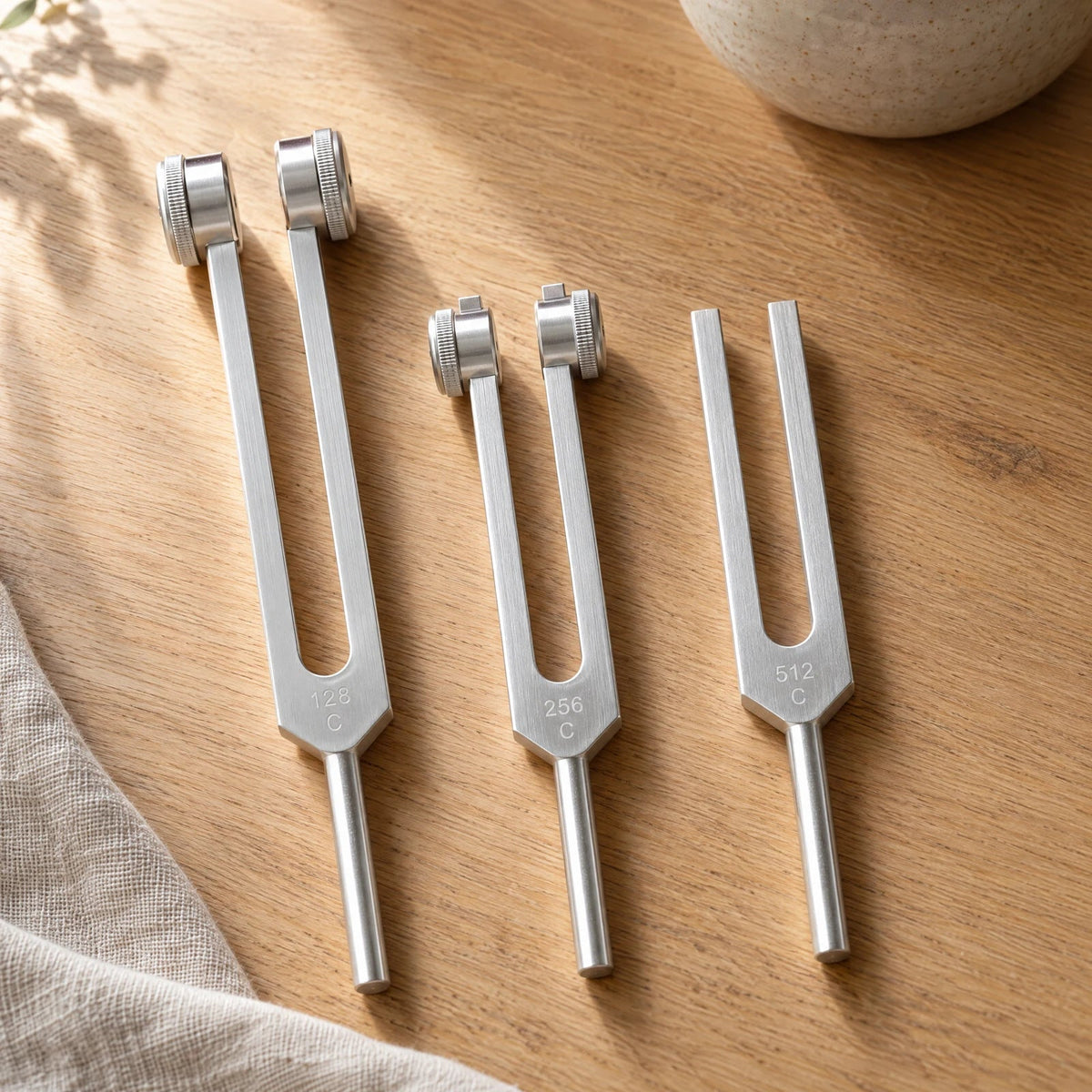 Three silver LotsOfZen tuning forks on a sunlit oak table beside ivory linen: 128 Hz and 256 Hz weighted forks, and a 512 Hz unweighted fork.