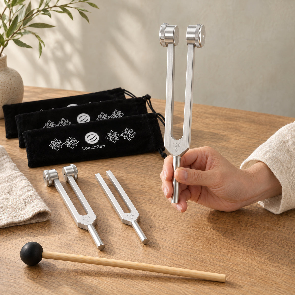 A hand holding the 128 Hz weighted fork above an oak table with the 256 and 512 Hz forks, wooden mallet and three velvet sleeves.