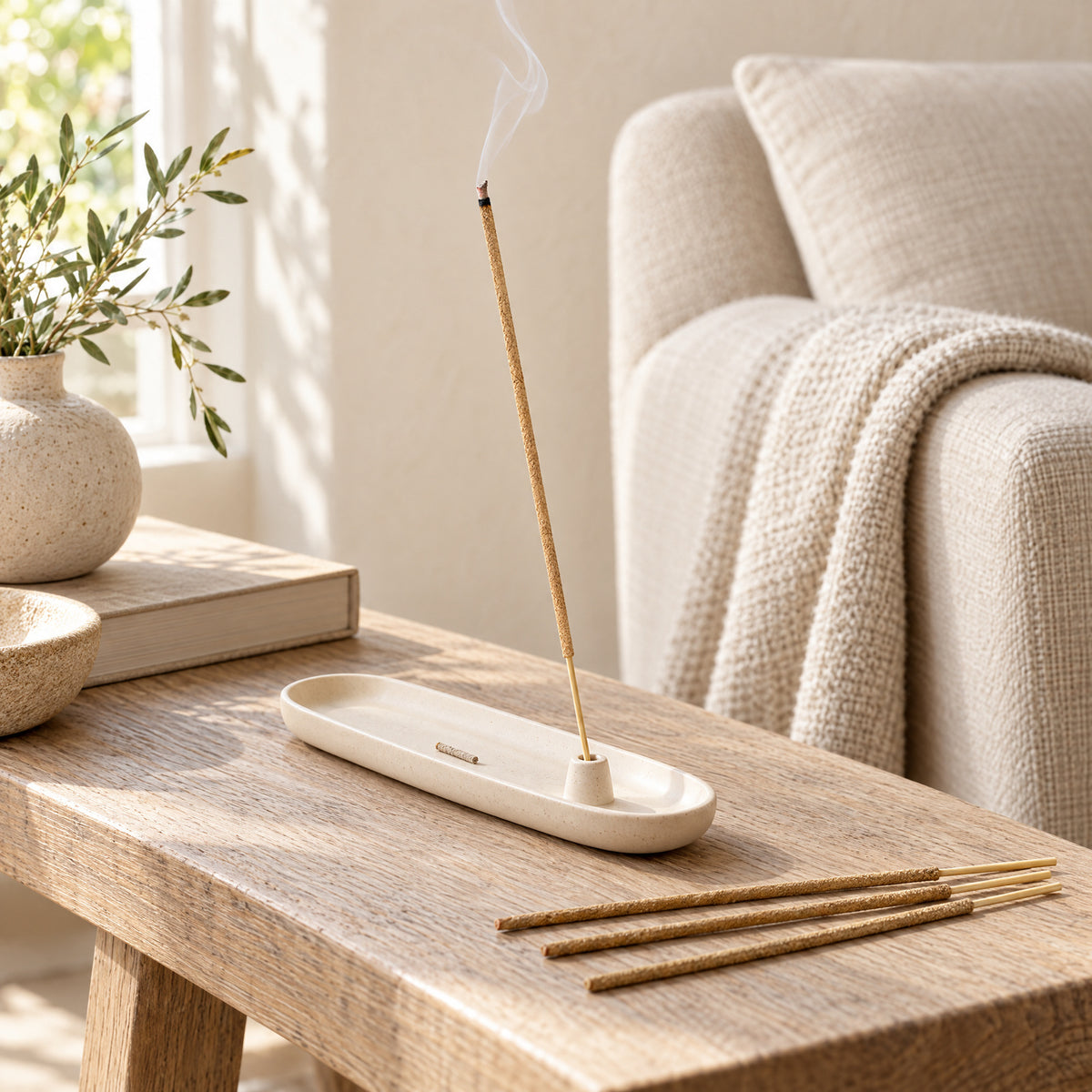 A Sandalwood incense stick smoldering in an ivory ceramic holder beside a linen chair.