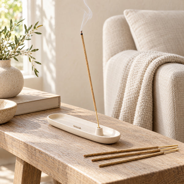 A Sandalwood incense stick smoldering in an ivory ceramic holder beside a linen chair.