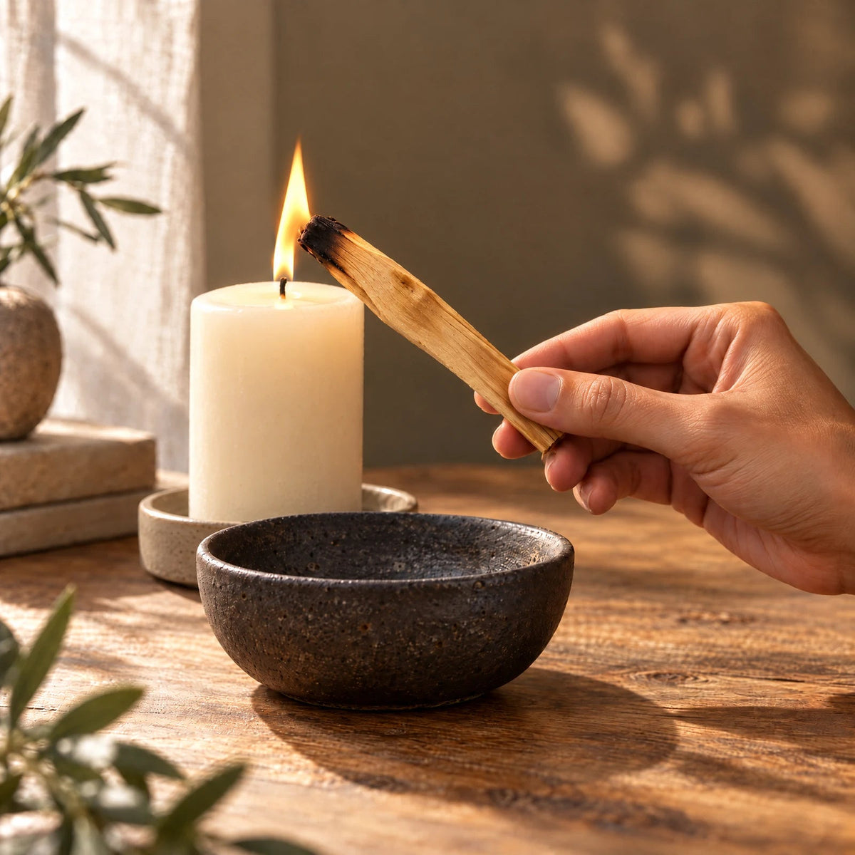 Styled scene of a hand holding Palo Santo by the clean end while its far tip touches a candle flame above a ceramic bowl