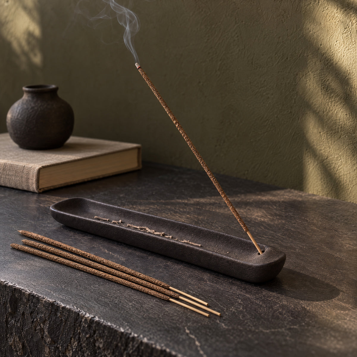 A brown Agarwood incense stick in a dark ash-catching tray on a slate console.