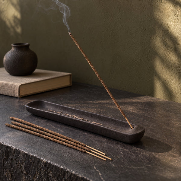 A brown Agarwood incense stick in a dark ash-catching tray on a slate console.