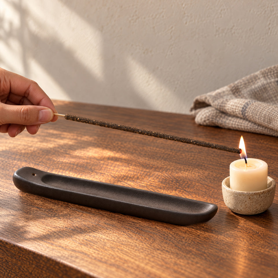 A hand holds the bare bamboo end while a candle lights the coated incense tip above an oak table.