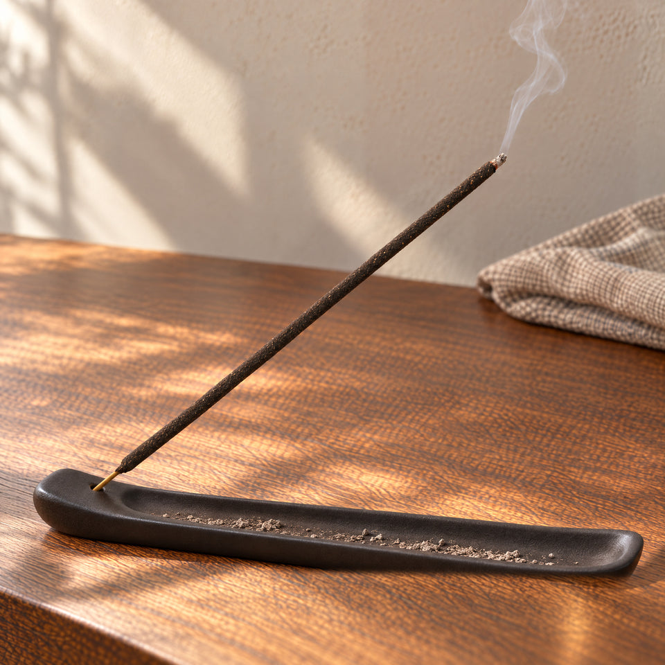 A smoldering bamboo incense stick rests securely in a dark ceramic holder, with fallen ash inside its tray.