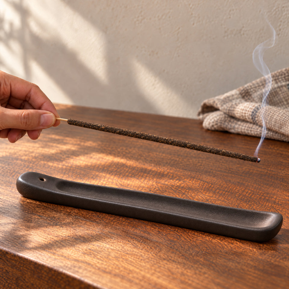 The same incense stick has a small glowing tip and a fine curl of smoke above a dark ash-catching tray.